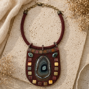 Statement bib necklace featuring deep garnet bead embroidery surrounding a dramatic druzy agate centerpiece, accented with earthy geometric beads, green cabochons, and antique brass findings.
