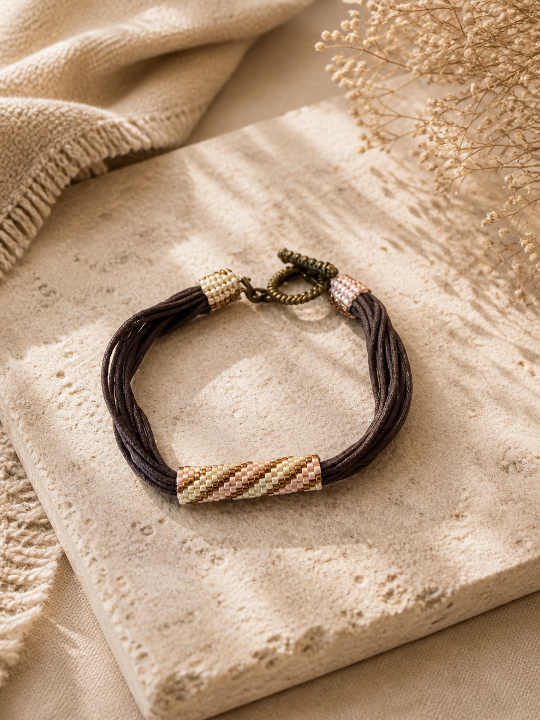 Multi-strand brown cord bracelet with soft pink and gold beadwoven accents and bronze toggle clasp.