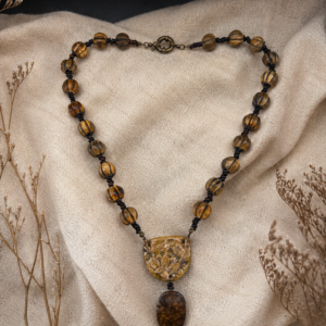 Earth-toned handmade necklace featuring amber glass beads, artisan ceramic focal, and dark lampwork pendant on a soft neutral fabric background.