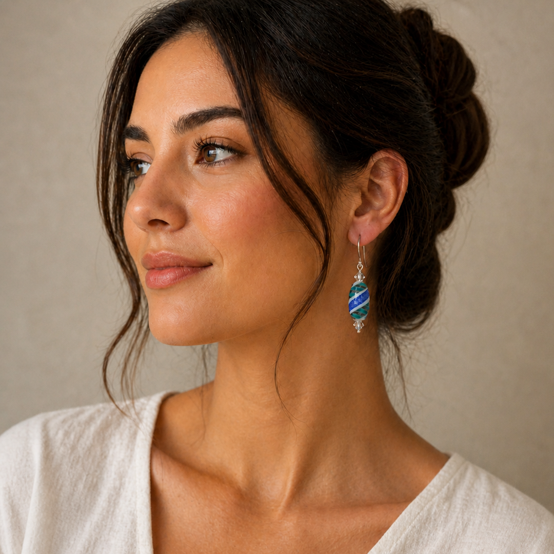 Portrait of a woman wearing handcrafted blue and turquoise lampwork-style drop earrings with sterling silver ear wires, styled against a soft neutral background in warm natural light.